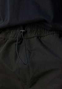 Zwarte elastische tailleband met trekkoord op zwarte stoffen broek, gecombineerd met een zwart shirt dat iets in de taille is gestopt.