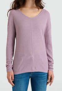 Světle fialový pletený svetr s výstřihem do V, žebrovanými manžetami a lemem. Má texturovaný vzor látky a uvolněný střih. Nošený s modrými džínami.