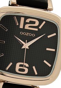 Montre noire avec un cadran carré, accents en or rose, chiffres 12 et 6 bien visibles, cadran noir texturé et un bracelet en cuir noir.