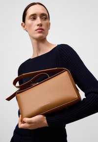 Borsa a tracolla in finta pelle marrone con forma rettangolare, lunga tracolla e un branding discreto sul davanti. Sfondo in maglione navy a maglia.