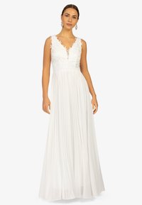 Robe blanche avec un décolleté en V profond, des accents en dentelle florale aux épaules et une jupe plissée. Le tissu a une texture douce et une drape élégante.