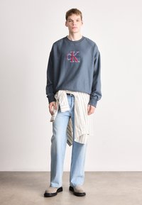 Sweatshirt azul claro com logo CK, combinado com uma camisa às riscas atada à cintura, jeans azul claro e calçado cinza com sola grossa.