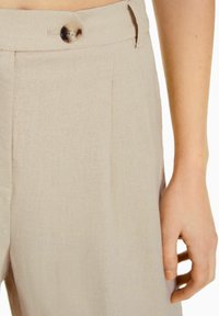 Beige högmidjade byxor med en enda knapp och bältesöglor, visade på en persons hand som vilar vid höfterna.