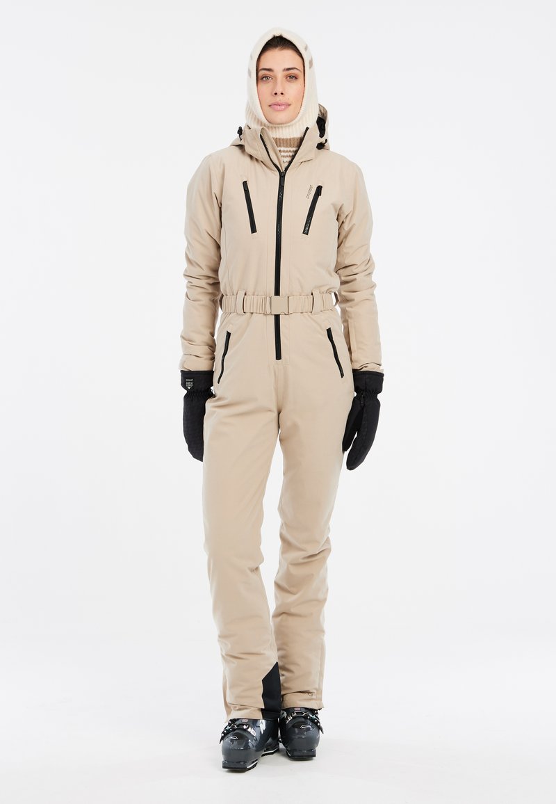 Completo da sci beige con vita cintata, caratterizzato da cerniere nere e polsini accentuati. Indossato con guanti neri e cappuccio lavorato a maglia. Materiale impermeabile.
