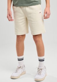 Bermudas de algodón beige con una cintura elástica, bolsillos laterales y un pequeño logo. Combinadas con zapatillas blancas y calcetines cortos blancos a rayas.