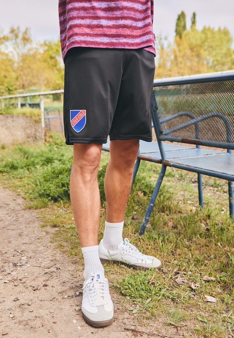 Fekete sportos rövidnadrág kék és piros emblémával, fehér zoknikkal és fehér sportcipőkkel párosítva. A rövidnadrág sima, könnyű anyagból készült.