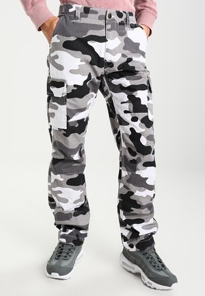 Person trägt schwarze, weiße und graue Camouflage-Cargohosen mit Seitentaschen und graue Sportschuhe, Hände in den Taschen.