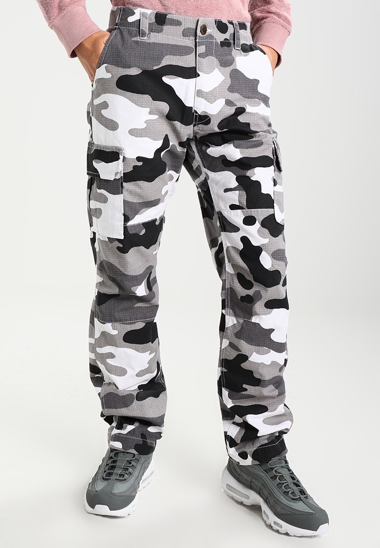 Personne portant un pantalon cargo camouflage noir, blanc et gris avec des poches latérales et des baskets athlétiques grises, mains dans les poches.