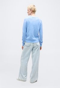 Άτομο με κοντά ξανθά μαλλιά που φοράει ανοιχτό μπλε πουλόβερ, ανοιχτό μπλε τζιν παντελόνι με φαρδιά μπατζάκια και λευκά παπούτσια, ποζάροντας με την πλάτη προς την κάμερα.