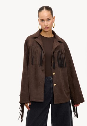 Veste en suède marron avec des franges sur le devant et les manches, dotée d'un col et de boutons dorés, accompagnée d'un jean foncé.