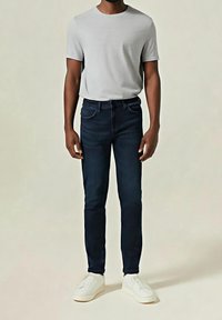 Vaqueros azul oscuro, diseño ajustado con estilo clásico de cinco bolsillos, combinados con una camiseta gris claro de cuello redondo y zapatillas blancas.