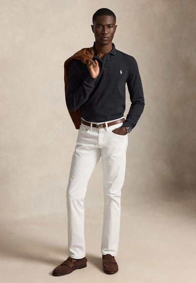 Vyras dėvi tamsų ilgomis rankovėmis polo marškinėlį, baltas kelnes su rudu diržu, rudas loaferius, laiko rudą švarką ant peties, stovi priešais smėlio spalvos foną.