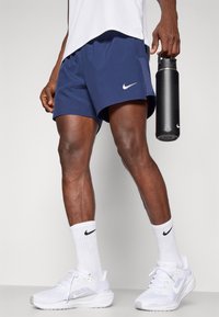 Granatowe spodenki sportowe z logo Nike, połączone z białymi skarpetkami z dzianiny i białymi butami Nike. Osoba trzyma czarną butelkę na wodę.