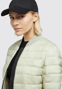 Light green puffer jacket with horizontal quilted design, ribbed collar, and black zipper; paired with a black baseball cap.