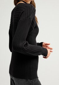 Zwarte geribbelde gebreide trui met een getailleerde silhouet, brede mouwen en textuur met verticale lijnen, gecombineerd met glinsterende donkere broeken.