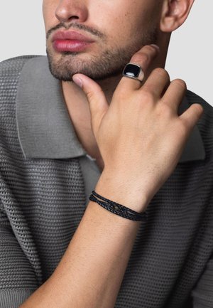 Black ring with a square onyx stone, worn on a hand with a braided black bracelet, against a textured gray polo shirt.
