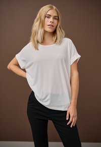 Blondīne ar plecu garuma matiem, valkājot vaļīgu baltu īss piedurkņu topu un melnas bikses, stāv pret brūnu fonu.