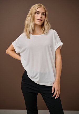 Õlgadeni blond juustega naine, kes kannab lahtist valget lühikeste varrukatega pluusi ja musti pükse, seisab pruuni tausta ees.