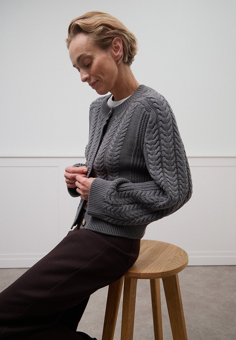 Grijze kabelgebreide cardigan met losse mouwen en ronde halslijn, gecombineerd met donkere broeken. De textuur is chunky; zittend op een houten kruk.