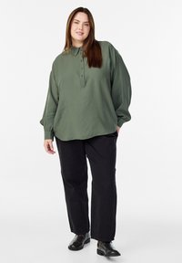 Kobieta stoi w oversize'owej oliwkowej koszuli zapinanej na guziki, czarnych spodniach i ciemnych skórzanych butach na tle białym.