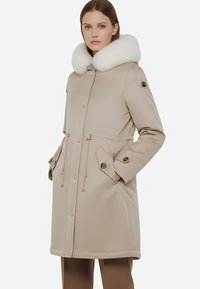 Cappotto impermeabile beige con colletto in ecopelliccia bianca, vita regolabile con cordino, polsini con bottoni e tasche frontali, con dettagli in hardware color oro.