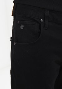 Black denim trousers feature a slim fit with five pockets. The fabric is smooth with visible stitching, and metallic buttons enhance the design.