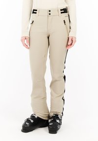 Beige skibroek met zwarte zijstrepen, ritszakken, en zwarte skischoenen met gespen bij een staand persoon.
