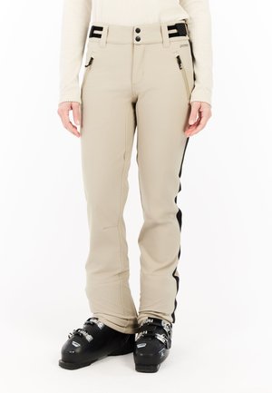 Beige Skihosen mit schwarzen Seitenstreifen, mit Reißverschlüssen versehenen Taschen und schwarzen Skischuhen mit Schnallen an einer stehenden Person.