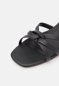 Sandale plate noire avec bout carré et trois brides en cuir torsadées à l'avant.