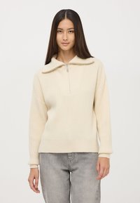 Pulower w kolorze kremowym, z prążkowanego dzianiny, z kołnierzem na pół zamku i dekoltem w kształcie litery V. Długie rękawy z dopasowanymi mankietami. Teksturowany materiał.