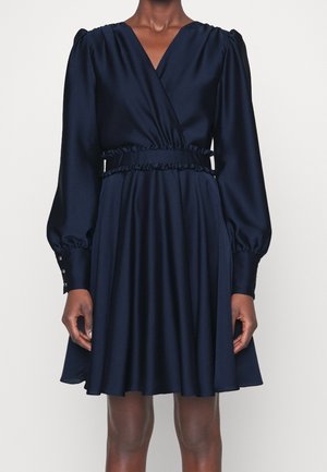 Vrouw draagt een marineblauwe wikkeljurk met lange mouwen, gerimpelde taille en knoopmanchetten, staand tegen een effen achtergrond.