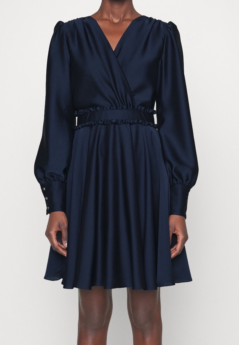 Femme portant une robe portefeuille bleu marine à manches longues avec des détails à volants à la taille et des poignets boutonnés, debout devant un fond uni.