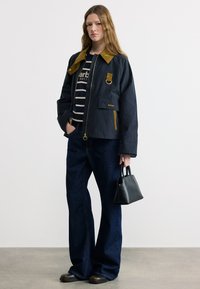 Νεαρή γυναίκα με μακριά μαλλιά, φοράει ναυτικό μπλε μπουφάν με καφέ γιακά, ριγέ πουκάμισο, μαύρο τζιν παντελόνι φαρδιάς γραμμής, μαύρα παπούτσια, κρατώντας μια μικρή μαύρη τσάντα.