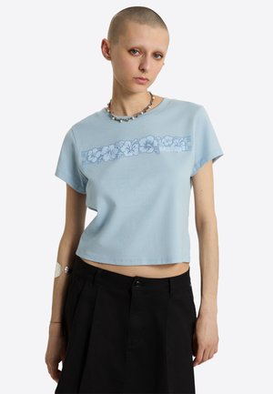 Jeune personne à la tête rasée portant un t-shirt court bleu clair avec un motif floral et un pantalon noir taille haute sur fond blanc.