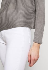 Pull gris côtelé avec une coupe décontractée, doté de manches longues et d'un ourlet tombant. Associé à un jean blanc, mettant en valeur une texture lisse.