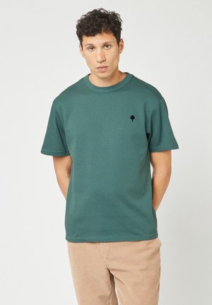 Jonge man met krullen, gekleed in een teal shirt met korte mouwen met een klein zwart boompje-logo en lichtbruine corduroy broek, staand tegen een witte achtergrond.