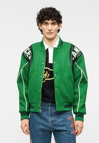 Πράσινο αθλητικό μπουφάν από μείγμα μαλλιού, με αντίθετα μαύρα δερμάτινα μανίκια και λευκή επένδυση. Κλείσιμο με κουμπιά και ριμπ μανσέτες.