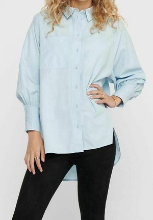 Femme portant une chemise boutonnée bleu clair oversize avec des manches bouffantes et un pantalon noir, posant avec une main sur la hanche.