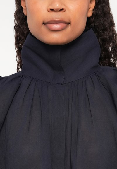 Blusa blu navy con collo alto e sottili plissettature, realizzata in tessuto leggero. Texture liscia con cuciture pulite e senza hardware visibile.