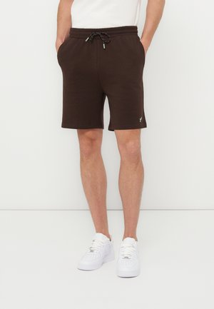 Mann som har på seg mørkebrune shorts med snor og hvite joggesko, står med hendene i lommene mot en ensfarget hvit bakgrunn.