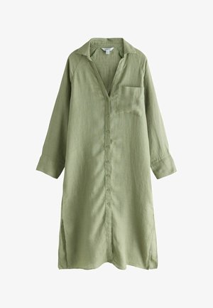 Femme en chemise en lin olive surdimensionnée, debout près d'un mur de roche texturé, regardant de côté avec l'océan et le ciel en arrière-plan.