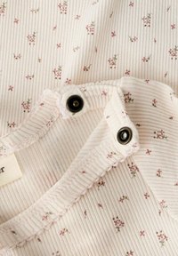 Close-up van een lichtgekleurde geribbelde stof met kleine roze bloemenprint en zwarte drukknoopjes op de halslijn van een babykledingstuk.