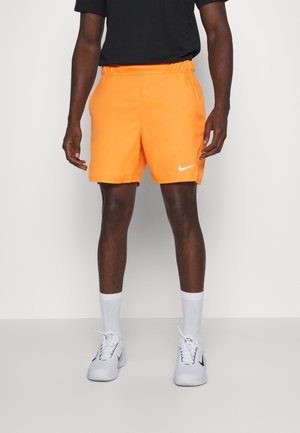 Homme portant un short Nike orange vif, des chaussettes blanches, des baskets Nike blanches et un t-shirt noir, debout devant un fond blanc uni.