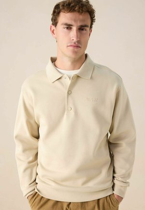 Beige polo shirt met lange mouwen en een gestructureerd oppervlak, knoopsluiting en subtiele borduurseldetail op de borst. Gecombineerd met corduroy broek.