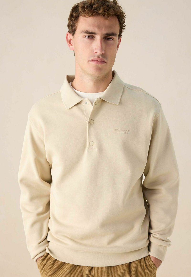 Polo à manches longues beige avec une surface texturée, patte de boutonnage et subtil détail de broderie sur la poitrine. Associé à un pantalon en velours côtelé.