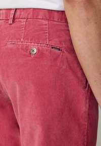 Pantalon en velours côtelé rose avec une texture à côtes verticales, une poche arrière avec fermeture à bouton et une petite étiquette de marque cousue.