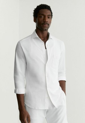 Mees seisab, üks käsi taskus, kandes tekstuuriga valget nupuga särki, mille varrukad on üles rullitud, ning sobivaid valgeid pükse.