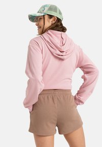 Un hoodie rose avec un cordon de serrage et une capuche, associé à un short beige clair. La casquette est verte avec un motif floral. Texture de tissu douce visible.