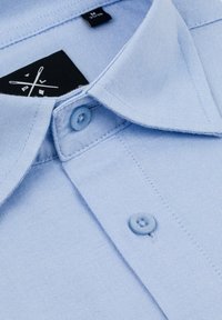 Jasnoniebieska koszula z długim rękawem z teksturowanej tkaniny. Posiada kołnierzyk, dwa guzik w pasującym kolorze oraz czarną metkę z logo wewnątrz kołnierza.