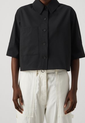 Persona che indossa una camicia nera corta con bottoni e taschino sul petto e pantaloni bianchi a vita alta con grandi tasche.